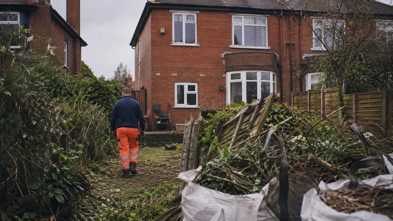 Garden clearance in progress on a 1930s red-brick semi-detached house with a bay window on a Wirral suburban street, coastal light